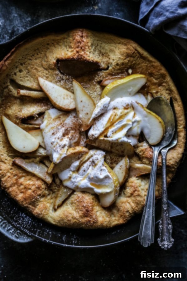 Autumn Pear Dutch Baby 6 A Puffed Pear Pancake, partially sliced, showing the soft interior and the juicy pear pieces. It is garnished with extra pears, whipped cream, and syrup.