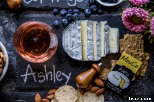 MouCo Cheese And Wine Pairing 5 A perfectly sliced portion of MouCo Ashley cheese, featuring its distinctive edible ash layer and soft white interior, accompanied by a glass of sparkling rosé wine.