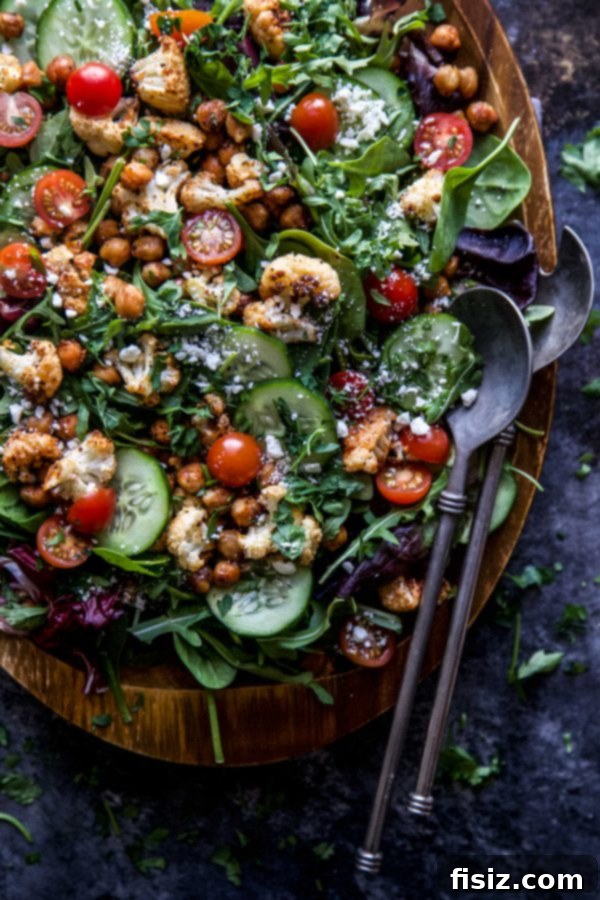 A close-up view of the roasted cauliflower and chickpeas being tossed with the fresh mixed greens, cucumbers, and tomatoes in a large salad bowl.