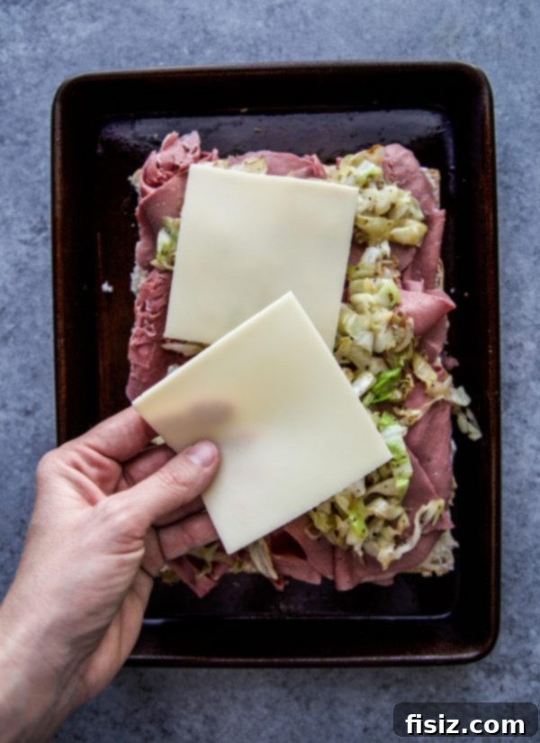 One-Pan Wonder Corned Beef Sliders 5 Hands carefully placing slices of Swiss cheese on top of the corned beef layers of the sliders.