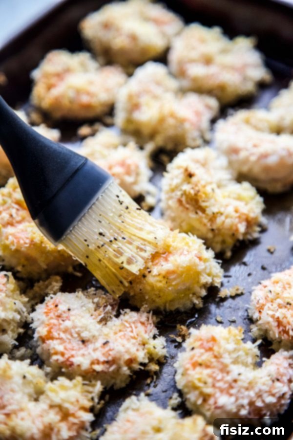 A chef's hand brushing golden hot honey sauce onto baked coconut shrimp on a baking sheet.