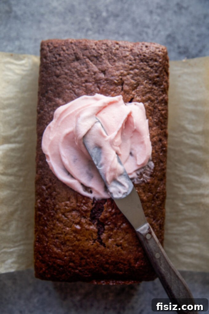 A chocolate loaf cake being frosted with a vibrant cherry frosting using a spatula.