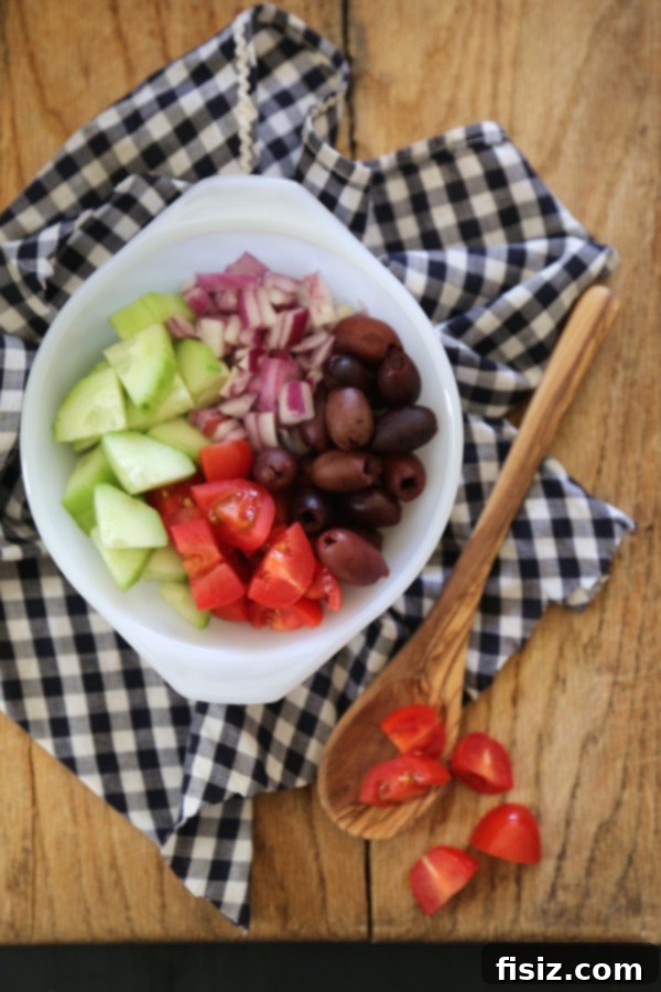 One-Pan Greek Feta Gnocchi 3 A white bowl showcasing fresh, colorful ingredients for Greek Feta Gnocchi Skillet, including halved grape tomatoes, thinly sliced red onion, diced cucumbers, and a mix of olives, with a wooden spoon nearby.